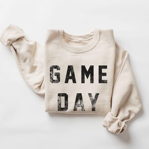 May include: A cream-colored sweatshirt with the words "GAME DAY" printed in distressed black lettering. The sleeves are folded, and the garment is laid flat on a white surface. A casual, comfortable style.