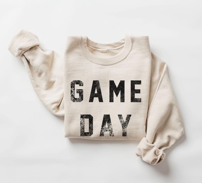 May include: A cream-colored sweatshirt with the words "GAME DAY" printed in distressed black lettering. The sleeves are folded, and the garment is laid flat on a white surface. A casual, comfortable style.