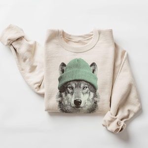 Puede incluir: Sudadera de color crema con un gráfico detallado de un lobo con un gorro verde. La cara del lobo está centrada, con el gorro cubriendo sus orejas. Las mangas están enrolladas y la sudadera está doblada cuidadosamente.