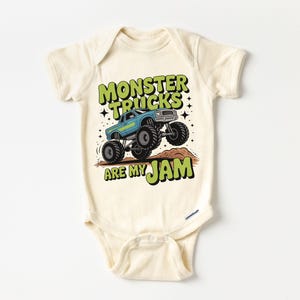 Puede incluir: Body de beb&eacute; color crema con un gr&aacute;fico de un cami&oacute;n monstruo azul saltando sobre un mont&iacute;culo marr&oacute;n. El texto "MONSTER TRUCKS ARE MY JAM" est&aacute; en verde y negro. El body tiene mangas cortas y cierres de presi&oacute;n.