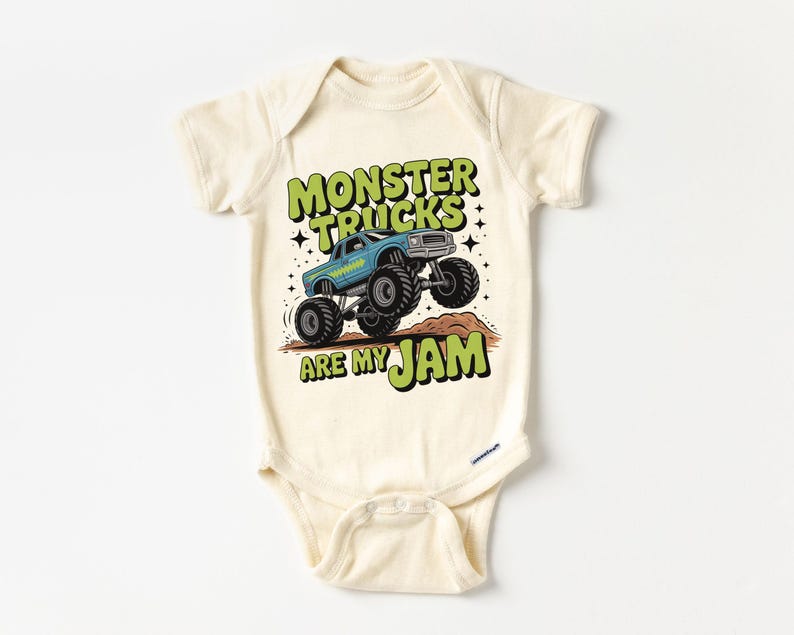 Puede incluir: Body de beb&eacute; color crema con un gr&aacute;fico de un cami&oacute;n monstruo azul saltando sobre un mont&iacute;culo marr&oacute;n. El texto "MONSTER TRUCKS ARE MY JAM" est&aacute; en verde y negro. El body tiene mangas cortas y cierres de presi&oacute;n.