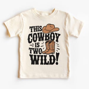 May include: Cream-colored toddler t-shirt with the text "THIS COWBOY IS TWO WILD!" in black. The design includes a brown cowboy hat and boots. A fun and playful design for a child's second birthday.