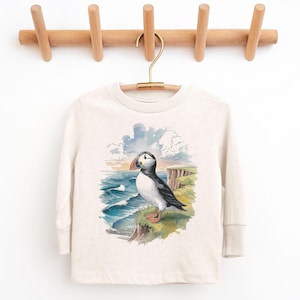 May include: A long-sleeved, light beige shirt featuring a watercolor illustration of a puffin bird perched on a cliff overlooking the ocean. The artwork includes blue waves, a cloudy sky, and green grass.