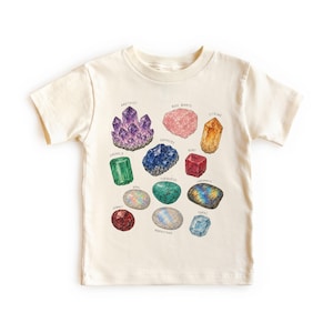 May include: A cream-colored short-sleeve t-shirt featuring a colorful graphic illustration of various gemstones including amethyst, rose quartz, citrine, emerald, sapphire, ruby, opal, turquoise, labradorite, garnet, moonstone, and topaz with labels.