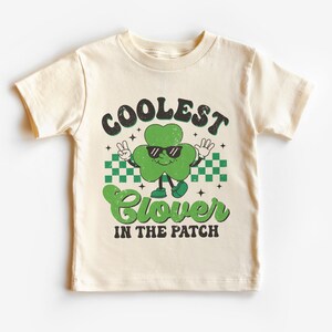 May include: A cream colored t-shirt with a green shamrock wearing sunglasses and the text "Coolest Clover in the Patch".
