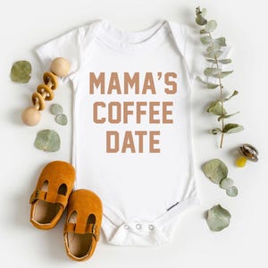 Puede incluir: Un body blanco de manga corta para beb&eacute; con el texto MAMA'S COFFEE DATE en letras marrones. La prenda est&aacute; rodeada por un sonajero de madera, un par de zapatos de cuero color amarillo mostaza, un chupete y hojas de eucalipto verdes.