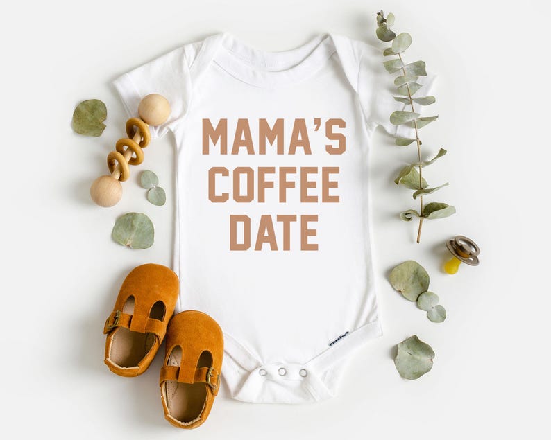 Puede incluir: Un body blanco de manga corta para beb&eacute; con el texto MAMA'S COFFEE DATE en letras marrones. La prenda est&aacute; rodeada por un sonajero de madera, un par de zapatos de cuero color amarillo mostaza, un chupete y hojas de eucalipto verdes.