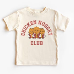 Pode incluir: Camiseta creme com as palavras "CHICKEN NUGGET CLUB" em um design de arco retrô. Abaixo do texto, três desenhos animados de pepitas de frango sorridentes usam jaquetas vermelhas. A camisa é de manga curta e gola redonda.