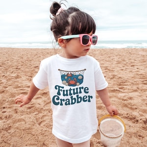 May include: A white t-shirt with the words "Future Crabber" in teal script. The shirt features a graphic of a crab, seashells, and waves inside a net. The child is wearing pink sunglasses and standing on a sandy beach.