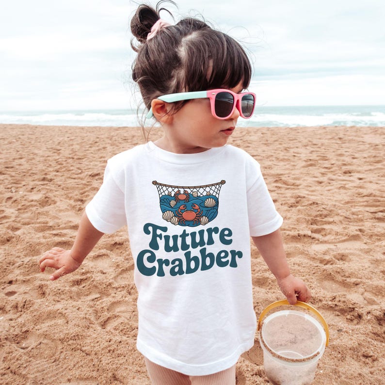 May include: A white t-shirt with the words "Future Crabber" in teal script. The shirt features a graphic of a crab, seashells, and waves inside a net. The child is wearing pink sunglasses and standing on a sandy beach.