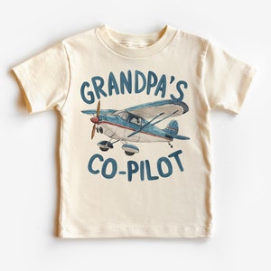 May include: A cream coloured t-shirt with a blue and white aeroplane graphic and the text "Grandpa's Co-Pilot" in blue.