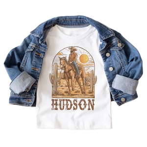 May include: White t-shirt with a graphic of a cowboy riding a horse in a desert landscape with cacti. The text "HUDSON" is printed below the graphic.
