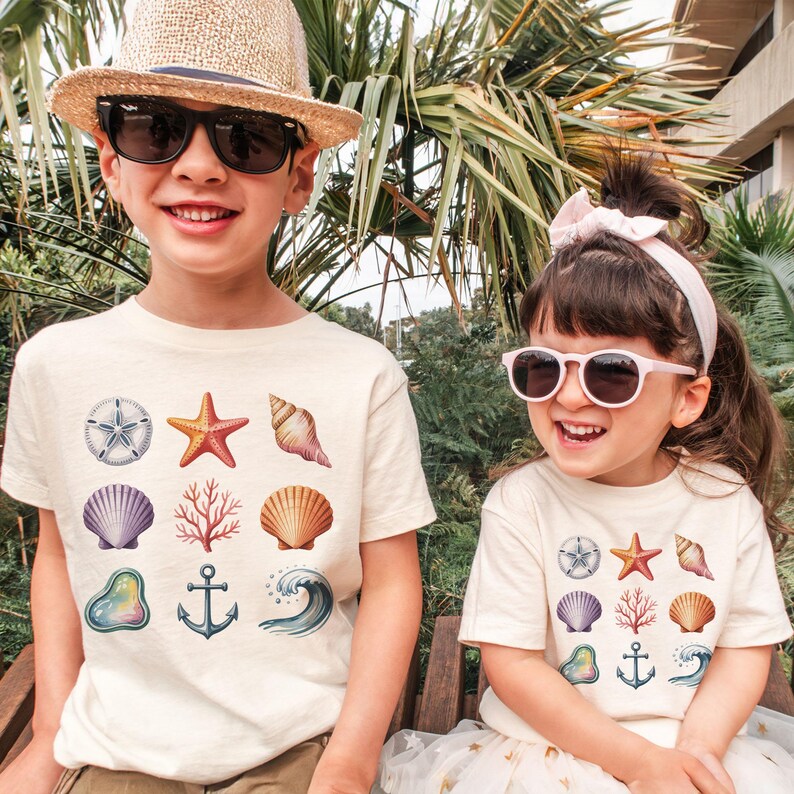 May include: Two children wearing cream-colored t-shirts with a colorful seashell graphic. The graphic includes a starfish, seashells, coral, and an anchor. One child wears a straw hat and sunglasses, the other wears a pink headband and sunglasses.