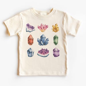 May include: Cream-colored t-shirt featuring a watercolor design of nine colorful crystals. The crystals are in shades of purple, pink, orange, blue, and green. The shirt is made of soft material and has short sleeves.