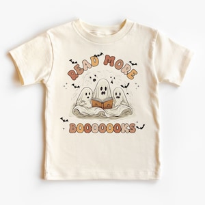 Op de afbeelding: Crèmekleurig T-shirt met korte mouwen met een Halloween-thema. Het ontwerp toont drie cartoongeesten die een boek lezen, met de woorden "READ MORE BOOOOOOKS" in oranje en bruine letters, en kleine vleermuizen.