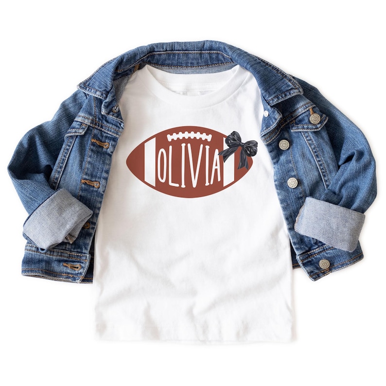 Puede incluir: Camiseta blanca con un dise&ntilde;o de bal&oacute;n de f&uacute;tbol americano marr&oacute;n y el nombre "OLIVIA" en letras blancas. Un lazo negro est&aacute; a la derecha. Una chaqueta vaquera azul est&aacute; encima.