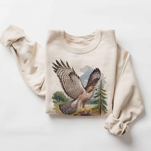 May include: A beige sweatshirt featuring a vintage illustration of a hawk in flight. The hawk is brown and white with a yellow beak and talons. The illustration is set against a green and blue background.