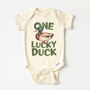 Puede incluir: Body de beb&eacute; color crema con el texto "ONE LUCKY DUCK" en letras verdes. Una ilustraci&oacute;n en acuarela de un pato con plumas verdes, marrones y rojas est&aacute; encima del texto. El body tiene mangas cortas y cierres de presi&oacute;n.