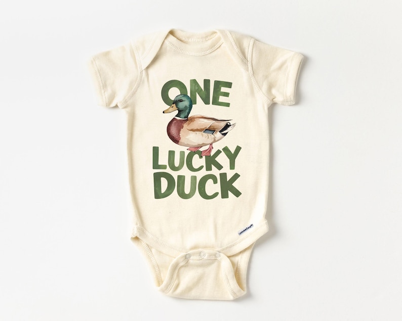Puede incluir: Body de beb&eacute; color crema con el texto "ONE LUCKY DUCK" en letras verdes. Una ilustraci&oacute;n en acuarela de un pato con plumas verdes, marrones y rojas est&aacute; encima del texto. El body tiene mangas cortas y cierres de presi&oacute;n.