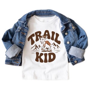 Peut inclure: T-shirt blanc avec un motif marron repr&eacute;sentant une ch&egrave;vre avec un sac &agrave; dos, le texte "TRAIL KID" et des silhouettes de montagnes. La chemise est associ&eacute;e &agrave; une veste en jean bleue. Un design amusant pour les amateurs de plein air.