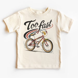 May include: A cream colored t-shirt with a graphic of a red and yellow bicycle with the words "Too fast" in black cursive lettering.