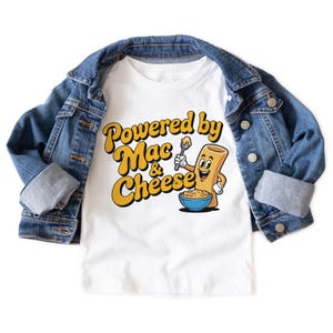K&ouml;nnte beinhalten: Wei&szlig;es T-Shirt mit einem Cartoon-Motiv einer l&auml;chelnden Makkaroni-Nudel, die einen L&ouml;ffel &uuml;ber einer Sch&uuml;ssel Makkaroni mit K&auml;se h&auml;lt. Der Text "Powered by Mac & Cheese" ist in Gelb und Orange gehalten. Eine blaue Jeansjacke ist &uuml;ber dem Shirt drapiert.