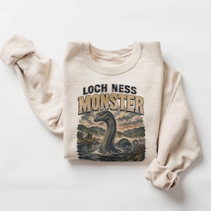May include: A cream-colored crewneck sweatshirt featuring a graphic print of the Loch Ness Monster. The text reads LOCH NESS MONSTER above an illustration of a long-necked creature emerging from dark water with mountains and a castle in the background.
