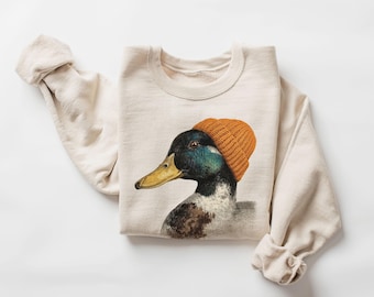 May include: Cream-colored sweatshirt featuring a graphic of a duck wearing an orange beanie. The duck has a green and black head, a yellow beak, and brown feathers. The sleeves are rolled up, and the sweatshirt is made of a soft material.