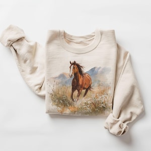 May include: A beige sweatshirt featuring a watercolor painting of a brown horse running through a field with mountains in the background. The horse has a white blaze and flowing mane. The sleeves are rolled up.