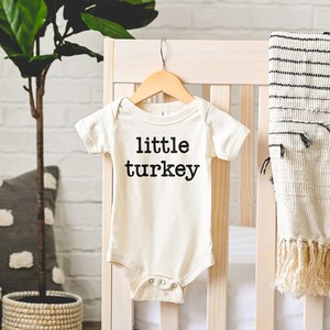 May include: Cream-colored baby bodysuit with the text "little turkey" in black. The short-sleeved onesie is hanging on a wooden hanger against a light-colored crib. A decorative blanket with fringe hangs on the crib.