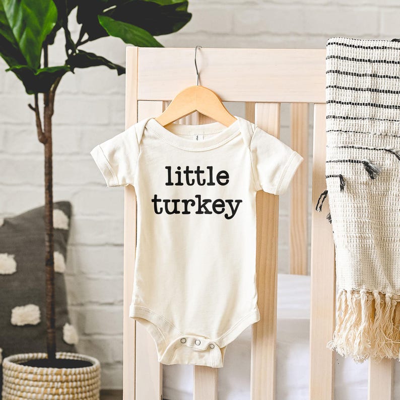 May include: Cream-colored baby bodysuit with the text "little turkey" in black. The short-sleeved onesie is hanging on a wooden hanger against a light-colored crib. A decorative blanket with fringe hangs on the crib.