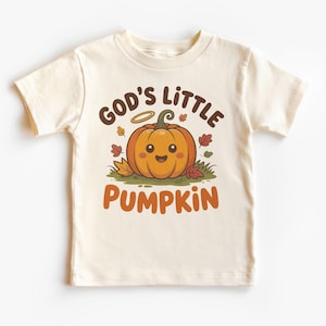 May include: A cream-colored short-sleeve t-shirt featuring a graphic of a smiling orange pumpkin with a halo. The text above reads "GOD'S LITTLE" in brown, and the word "PUMPKIN" in orange is printed below the pumpkin, surrounded by autumn leaves.