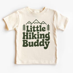 May include: Cream-colored t-shirt with the text "Little Hiking Buddy" in dark green. The design includes mountain and tree graphics above the text. The shirt is a short-sleeved crew neck.