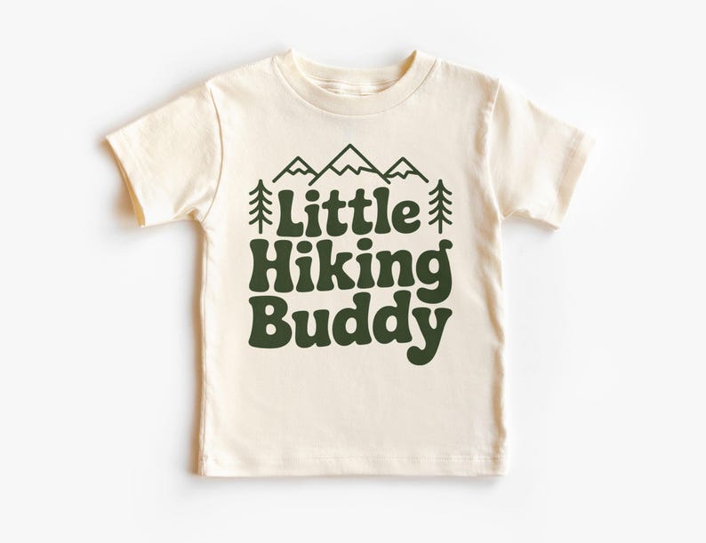 May include: Cream-colored t-shirt with the text "Little Hiking Buddy" in dark green. The design includes mountain and tree graphics above the text. The shirt is a short-sleeved crew neck.