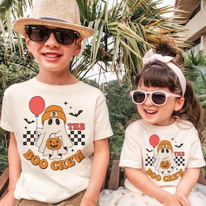 May include: Two children wearing matching cream-colored t-shirts with a Halloween-themed graphic. The graphic features a ghost holding a pumpkin and a red balloon, with the text "The Boo Crew". The shirts have a retro design with orange and black accents.