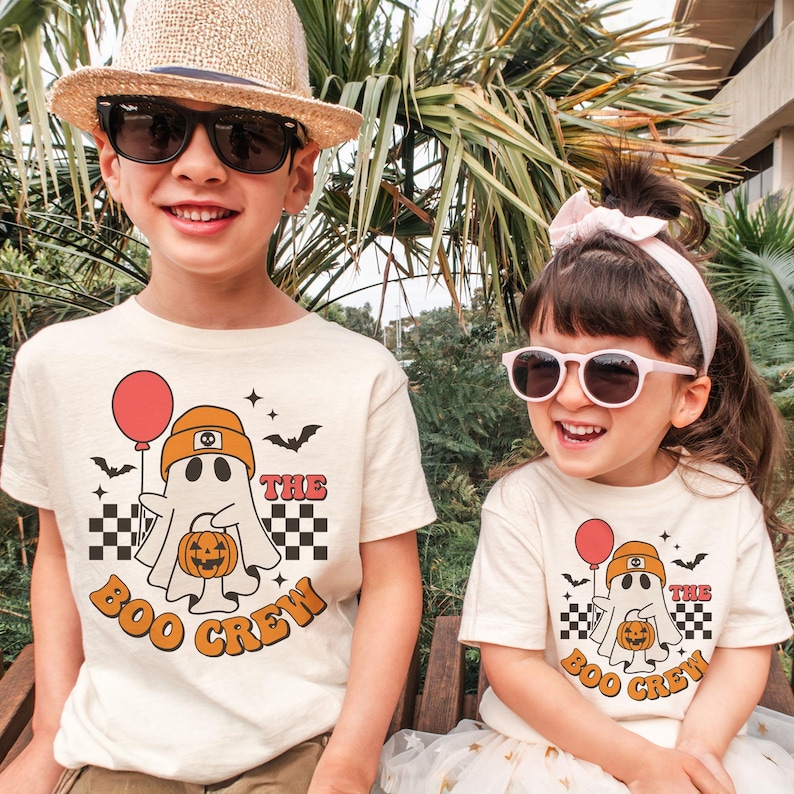 May include: Two children wearing matching cream-colored t-shirts with a Halloween-themed graphic. The graphic features a ghost holding a pumpkin and a red balloon, with the text "The Boo Crew". The shirts have a retro design with orange and black accents.