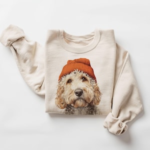 May include: Cream-colored sweatshirt featuring a dog wearing an orange beanie. The dog has light brown fur and a focused expression. The sleeves of the sweatshirt are rolled up. The overall aesthetic is casual and cozy.