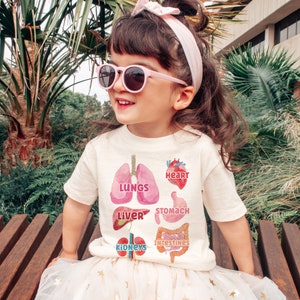 K&ouml;nnte beinhalten: Ein wei&szlig;es T-Shirt mit einer bunten Illustration der menschlichen Organe, darunter Lunge, Herz, Leber, Magen, Darm und Nieren. Der Text "LUNGS", "HEART", "LIVER", "STOMACH", "INTESTINES" und "KIDNEYS" ist auf dem Shirt aufgedruckt.