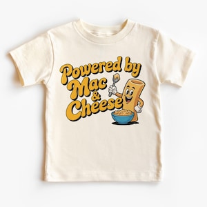 以下が含まれることがあります： クリーム色の子供用Tシャツ。黄色と黒で「Powered by Mac & Cheese」の文字。漫画のマカロニのキャラクターが、マカロニチーズのボウルの上にスプーンを持っています。
