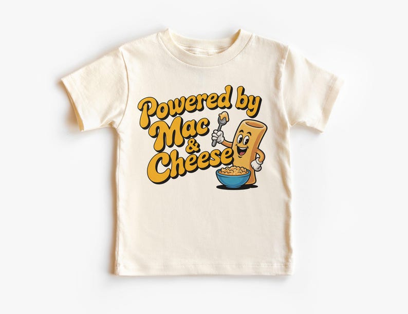 K&ouml;nnte beinhalten: Cremefarbenes Kinder-T-Shirt mit dem Text "Powered by Mac & Cheese" in Gelb und Schwarz. Eine Cartoon-Makkaroni-Figur h&auml;lt einen L&ouml;ffel &uuml;ber einer Sch&uuml;ssel Makkaroni mit K&auml;se.