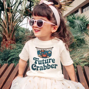 May include: A cream-colored t-shirt with a graphic of crabs, shells, and a net, with the words "Future Crabber" in teal. The child is wearing pink sunglasses and a matching headband.