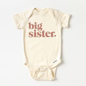 May include: A cream-colored baby onesie with short sleeves and the text "big sister" printed in a reddish-brown color.