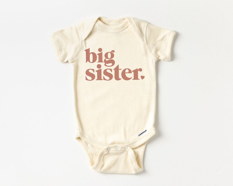 May include: A cream-colored baby onesie with short sleeves and the text "big sister" printed in a reddish-brown color.