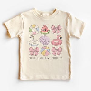 May include: A cream-colored t-shirt with a graphic of various colorful inflatable pool toys, including a watermelon, a unicorn, a flamingo, a seashell, a beach ball, and a life preserver. The text "Chillin with my floaties" is printed below the graphic.
