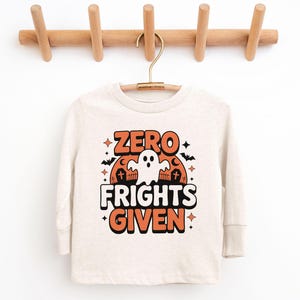 May include: A long-sleeved, off-white shirt with the text "ZERO FRIGHTS GIVEN" in orange and white, featuring a ghost, bats, and tombstones. The shirt is hanging on a wooden coat rack.