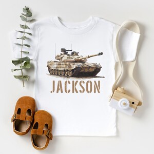 May include: White t-shirt with a camouflage tank printed on the front. The tank is brown and green with a white background. The name "JACKSON" is printed below the tank in brown letters.