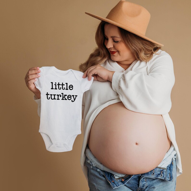 May include: A white baby onesie with the text "little turkey" in black. The onesie is held up in front of a pregnant person. The person is wearing a white blouse, blue jeans, and a tan hat. The background is a solid tan color.