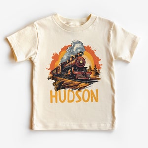 May include: A cream colored t-shirt with a vintage illustration of a train engine with smoke coming from the chimney. The train is traveling through a forest with a sunset in the background. The text "HUDSON" is printed in yellow below the train.