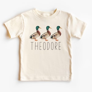 Może przedstawiać: Kremowa koszulka z nadrukiem trzech kaczek w rzędzie. Pod kaczkami znajduje się napis "THEODORE".