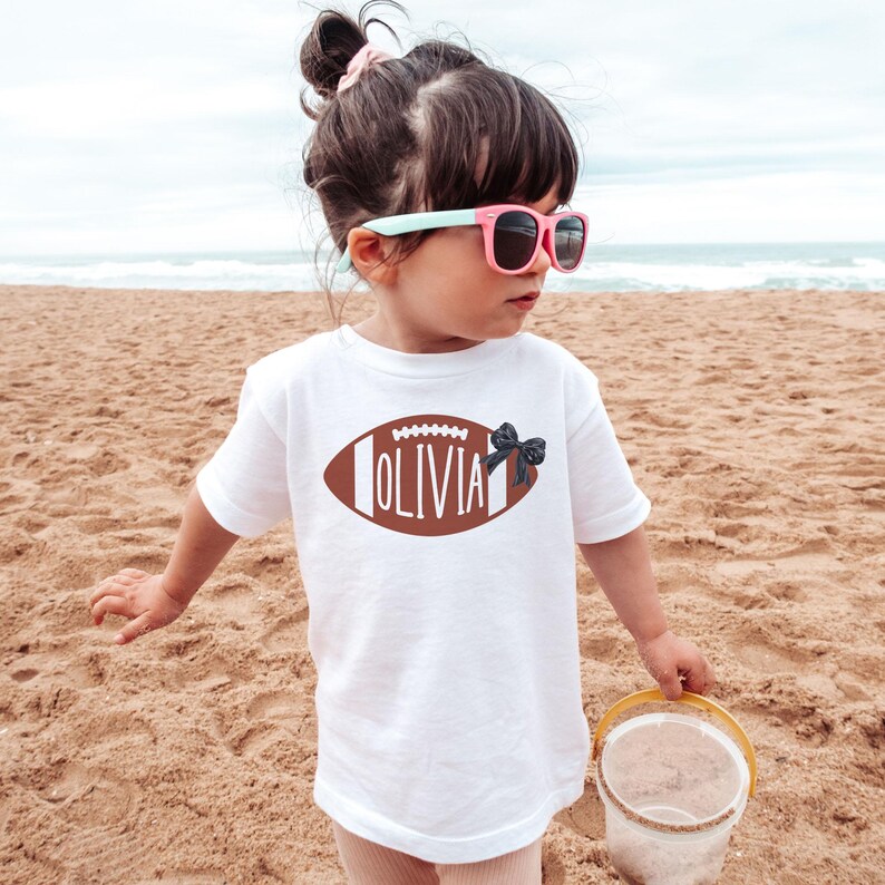 Puede incluir: Una camiseta blanca con un dise&ntilde;o de bal&oacute;n de f&uacute;tbol americano marr&oacute;n y el nombre "OLIVIA" en letras blancas. Un lazo negro est&aacute; en el lado derecho del bal&oacute;n. El ni&ntilde;o lleva gafas de sol rosas en una playa de arena.